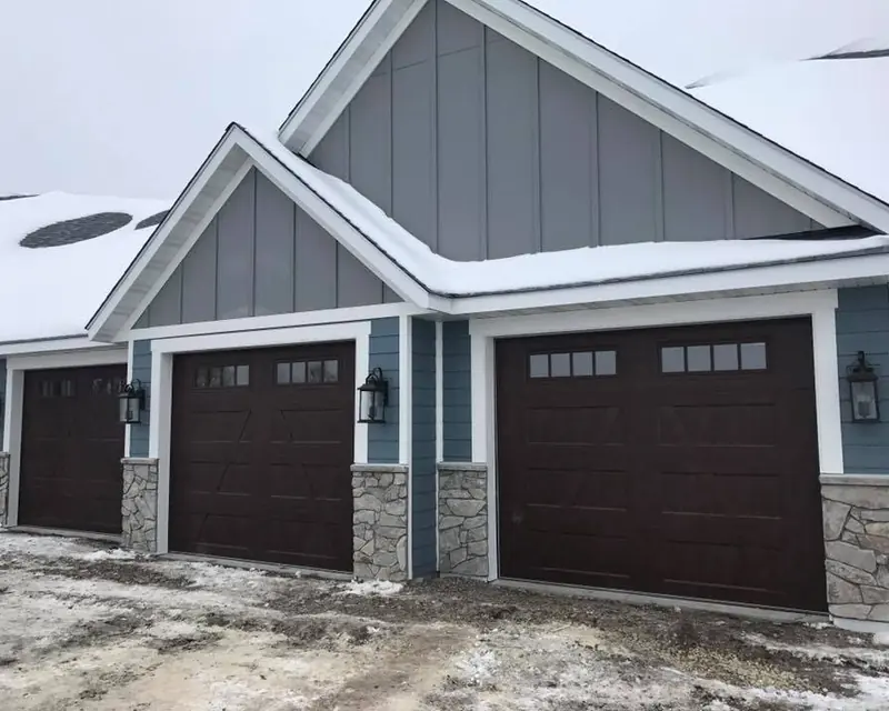 Three dark brown garage doors with upper windows on a blue-gray home